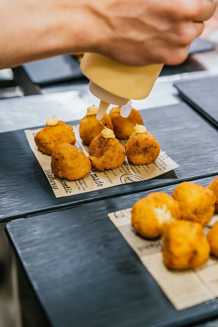 Anonymous chef hands meticulously drizzle a creamy sauce on golden croquettes arranged on a newspaper-lined serving tray