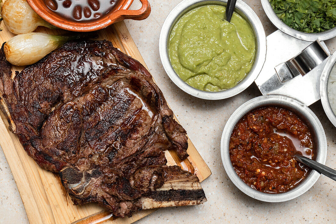 From above delectable Mexican-style grilled steak served on a wooden board, accompanied by traditional sides of beans, guacamole, salsa, and grilled onions, showcasing a well-seasoned and seared rib steak.