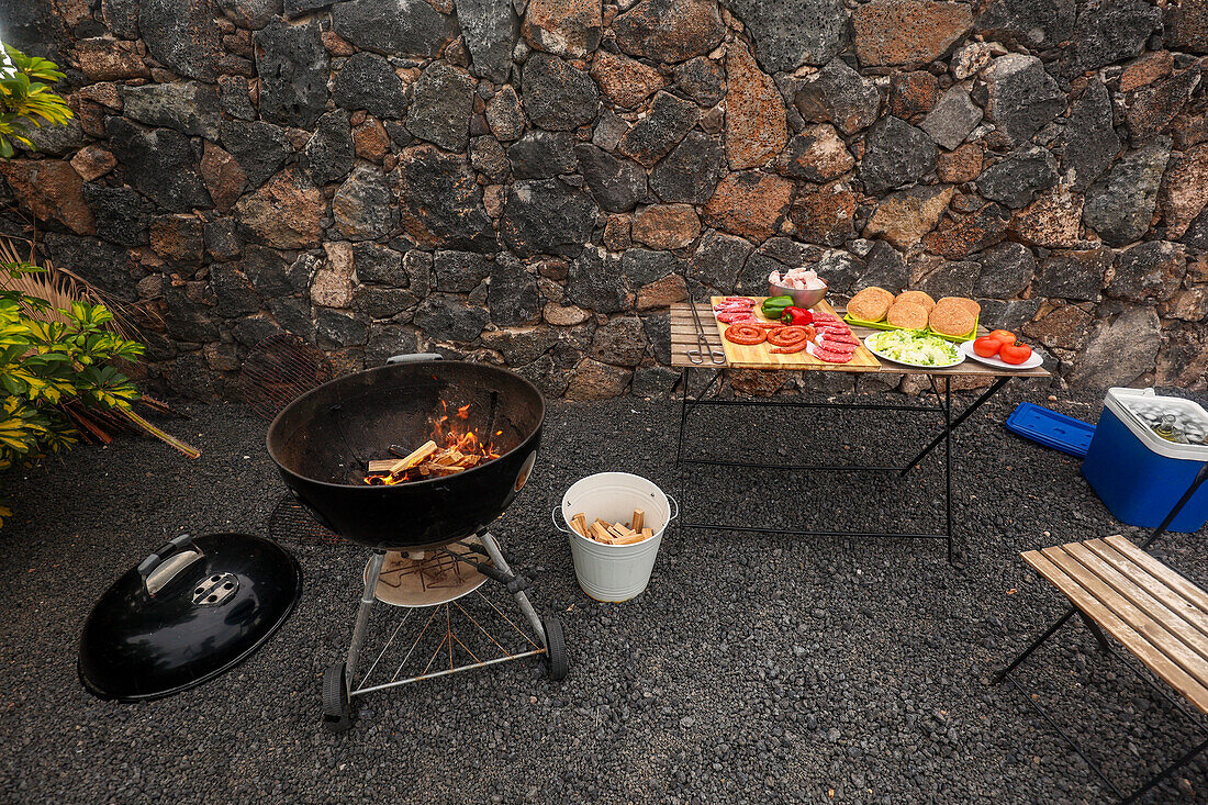 A charcoal grill actively burning, next to a table lined with sliced vegetables, meats, and buns for a barbecue setup outdoors, against a stone wall backdrop.