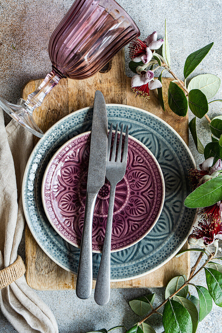 Draufsicht auf einen wunderschön gedeckten Tisch mit Tellern mit komplizierten Mustern in Violett- und Blautönen, ergänzt durch ein violettes Glas und mit zarten Feijoa-Blüten und -Blättern akzentuiert