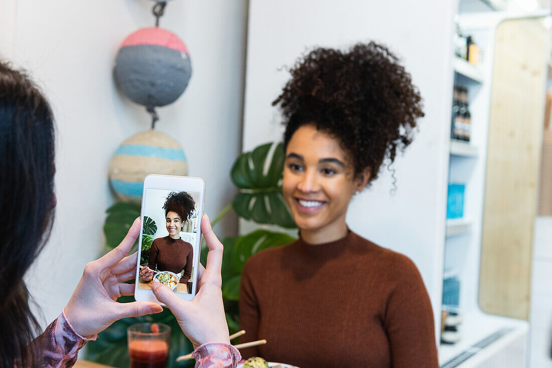 Eine nicht erkennbare weibliche Person fotografiert einen afroamerikanischen Freund, der in einem Restaurant mit einem Poke-Gericht am Tisch sitzt