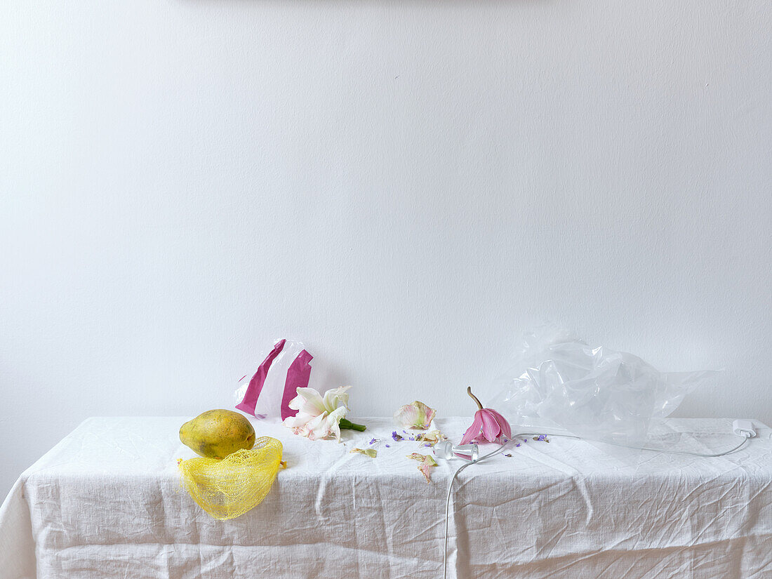 A gently composed still life image showcasing a fruit, scattered flower petals, a torn plastic bag, and a draped fabric table against a neutral background