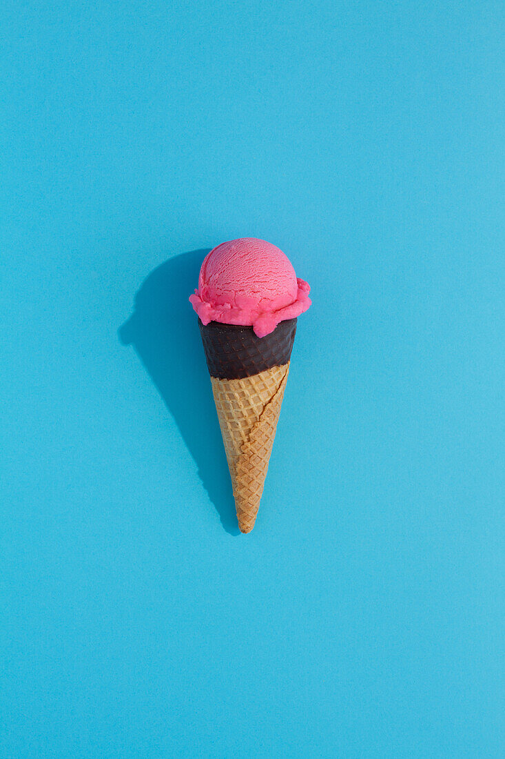 A vibrant Strawberry ice cream scoop sits atop a classic waffle cone against a blue background, casting a playful shadow