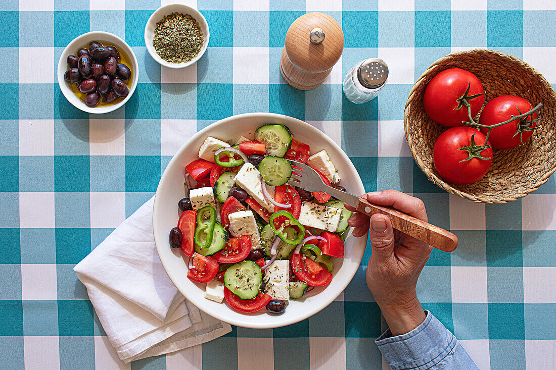 A fresh Greek salad served in a white bowl on a blue checkered tablecloth, featuring tomatoes, cucumbers, olives, and feta cheese, with a hand using a fork