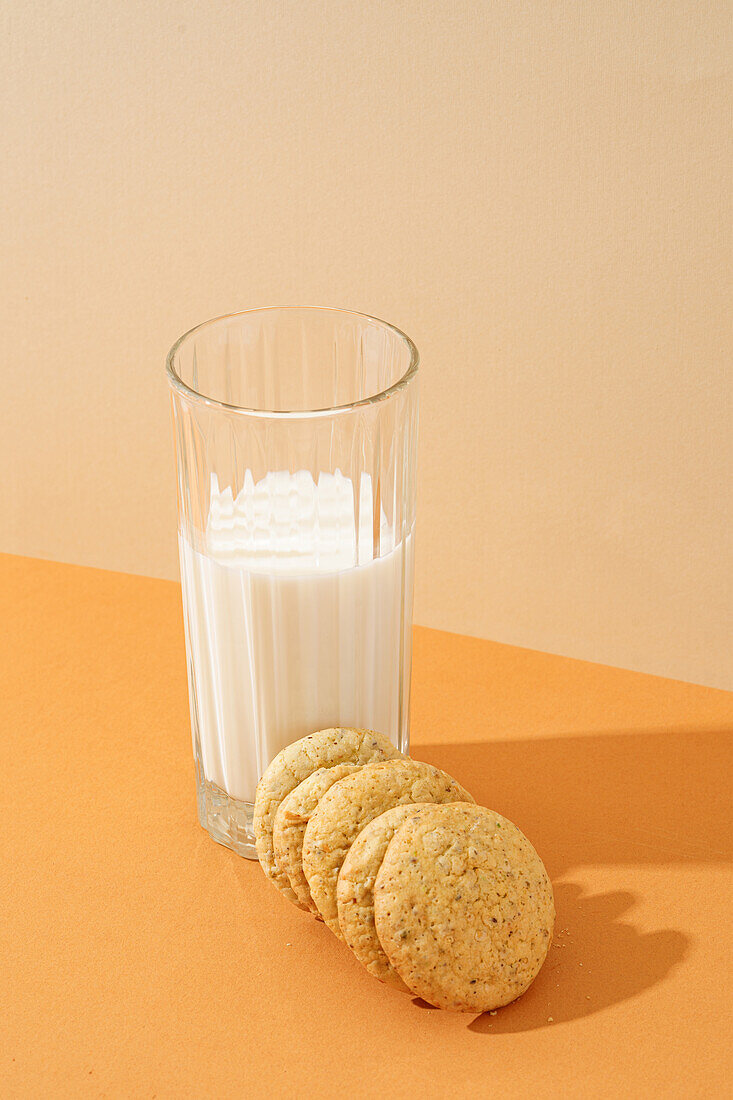 Ein ordentlicher Stapel knuspriger Haferflockenkekse neben einem vollen Glas Milch auf einer orangefarbenen Fläche, mit weichen Schatten.