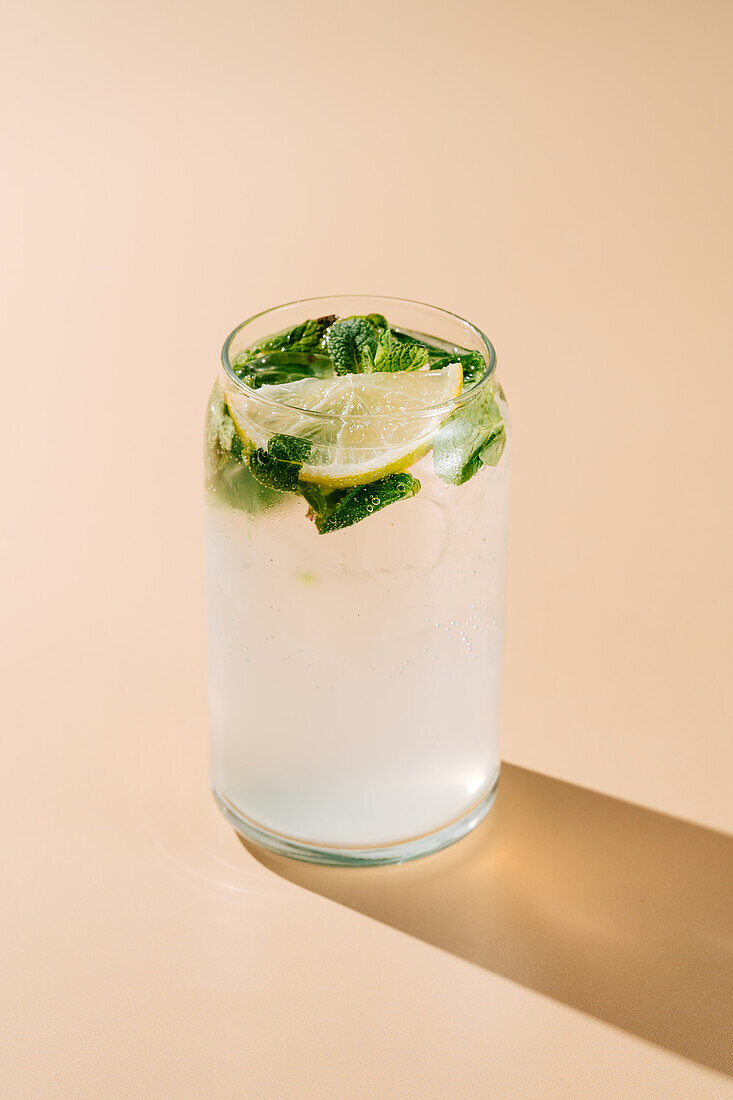 A classic mojito cocktail featuring fresh mint, slices of lemon, and bubbling soda water served in a glass, casting a shadow on a tan background.