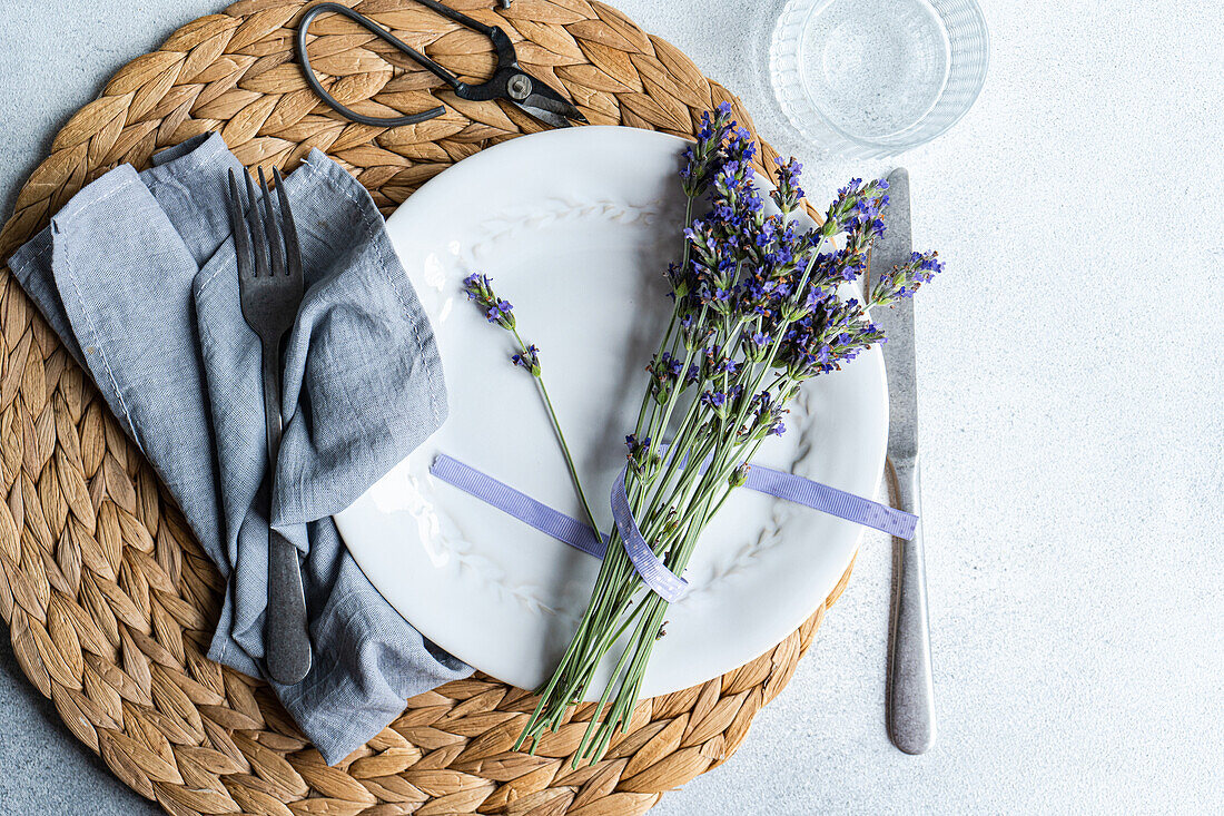 An elegant summer table setting featuring a white plate, grey linen napkin, and fresh lavender sprigs tied with a purple ribbon, all presented on a woven placemat