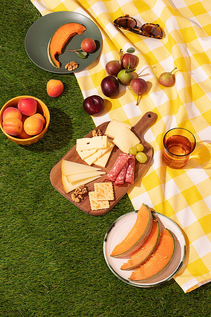 Top view of vibrant summer picnic scene on green grass, featuring a checkered yellow and white blanket with a selection of fresh fruits, cheeses, and snacks. A sunny day enhances the colorful setup.