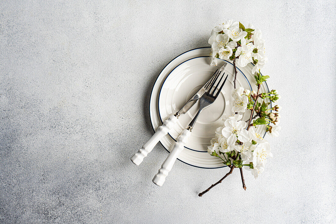 Elegant gedeckter Frühlingstisch mit weißen Blütenzweigen und Besteck im Vintage-Stil auf einem strukturierten grauen Hintergrund