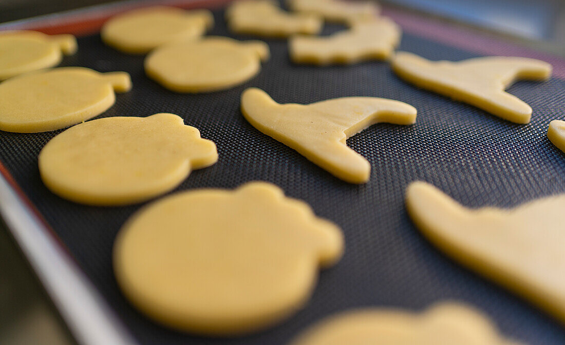 Eine Nahaufnahme von rohen Halloween-Keksen auf einem Backblech Die Formen umfassen Kürbisse und Hexenhüte, bereit zum Backen Der Schwerpunkt liegt auf der Textur des Teigs und den verwendeten festlichen Ausstechern