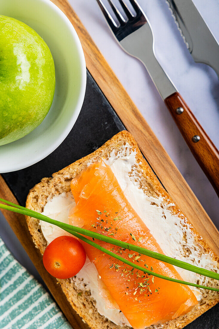 Top view showcasing a nutritious food arrangement featuring a vibrant apple, a sumptuous salmon sandwich on wholegrain bread layered with cream cheese, garnished with chives and a cherry tomato. Ideal for a wholesome meal theme.