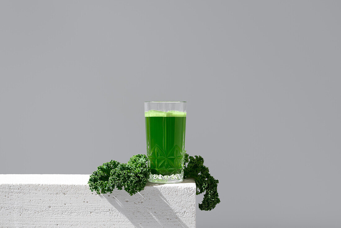 Ein Glas mit leuchtend grünem Grünkohlsaft, begleitet von frischen Grünkohlblättern auf einer weißen Fläche vor einem grauen Hintergrund