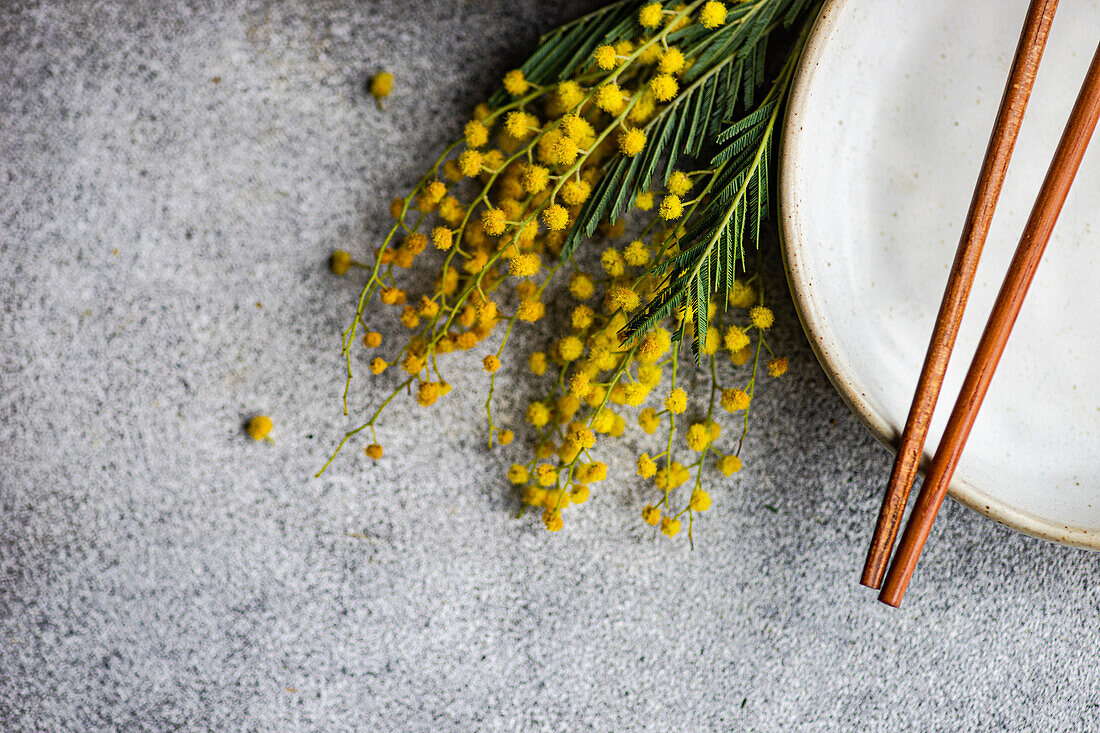 A detailed view of delicate mimosa flowers next to ceramic tableware with elegant, copper chopsticks on a textured grey surface