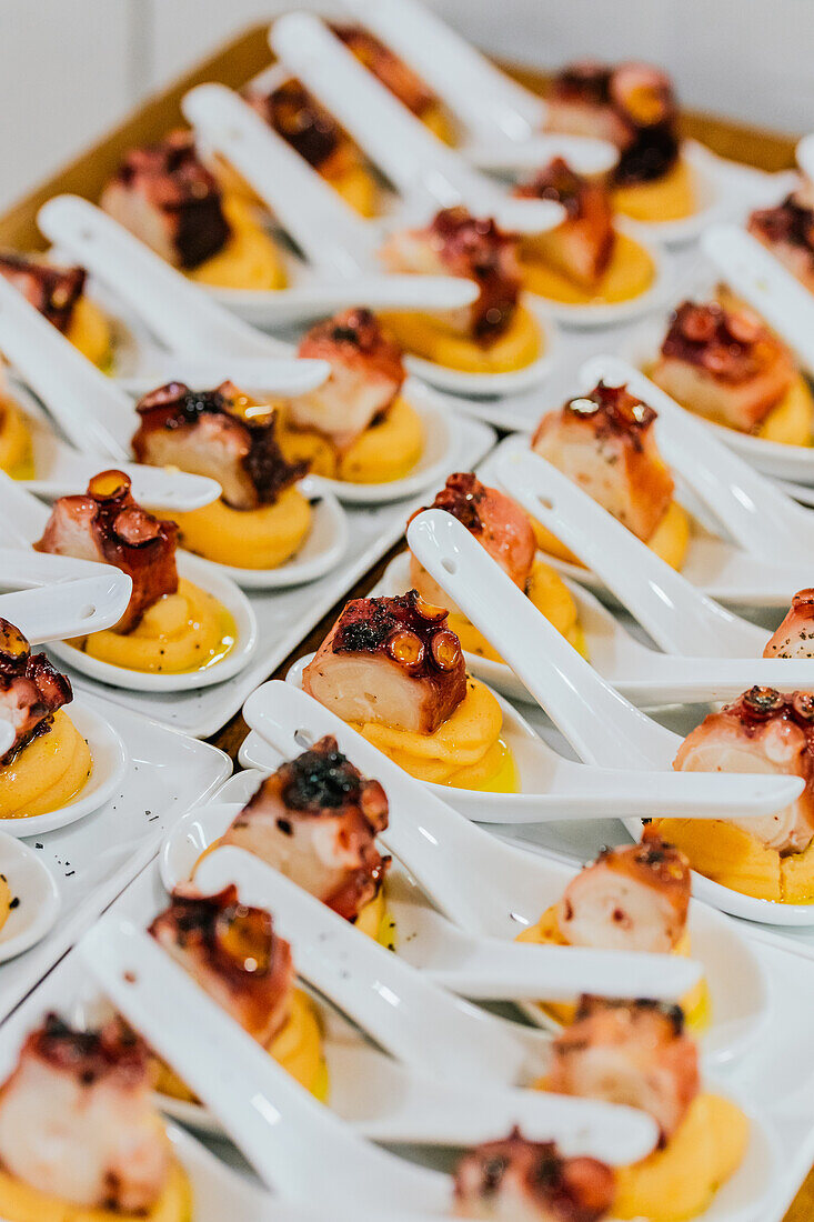 Rows of chic white spoons hold perfectly cooked octopus pieces atop a dollop of sauce, ready for a high-end gastronomic event