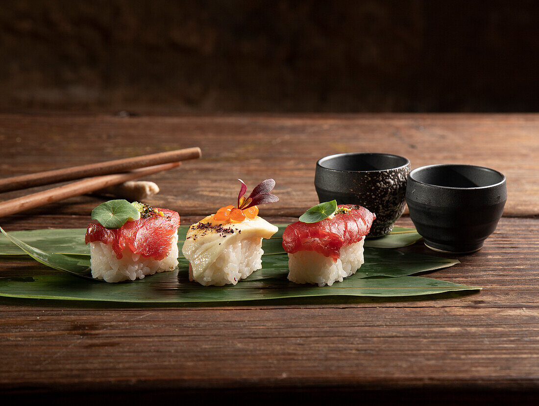 Ein Trio von Sushi-Stücken, die elegant auf einem breiten grünen Blatt präsentiert werden, begleitet von dunklen Keramikschalen und Essstäbchen, die die Essenz der japanischen Kochkunst einfangen