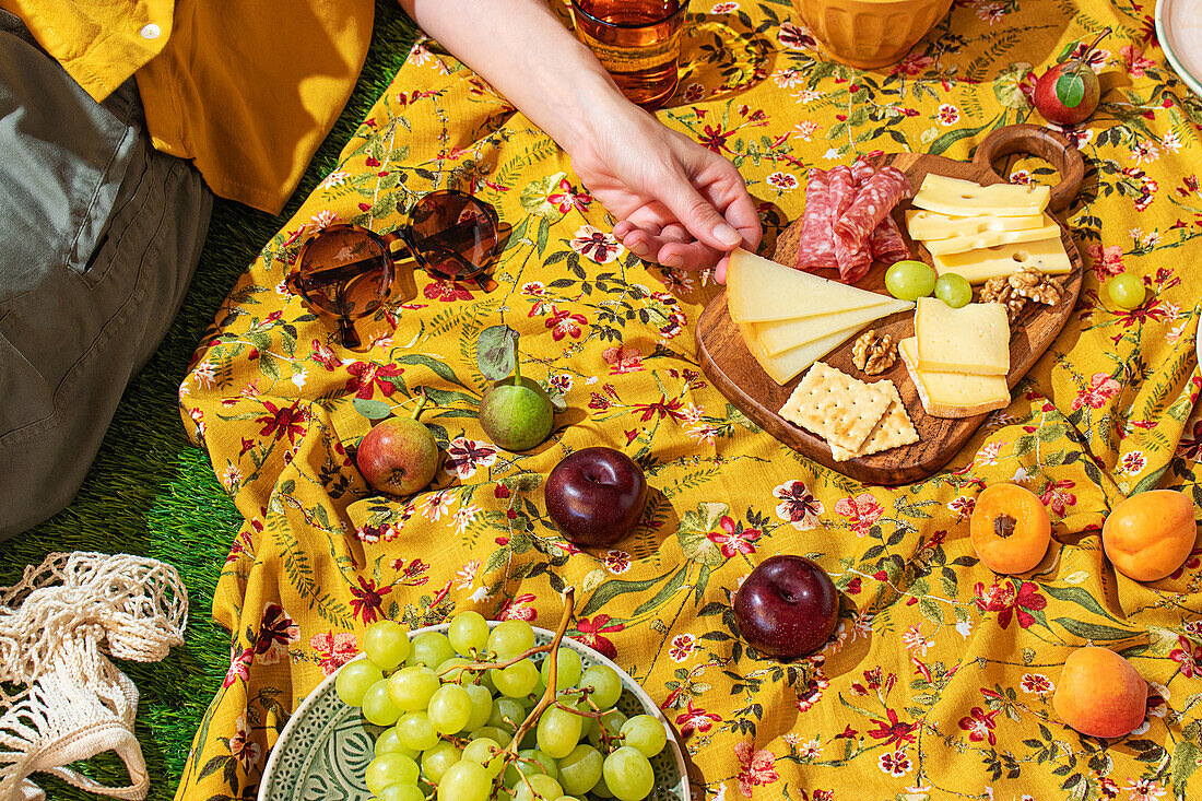 Detaillierte Szene eines Sommerpicknicks auf einer bunten, mit Blumen gemusterten Decke im Sonnenlicht. Eine Hand greift zwischen frischem Obst und Getränken nach Käse und Fleisch und fängt die entspannte Freude an einem Essen im Freien ein.