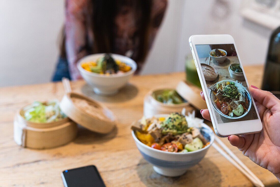 Cropped unrecognizable woman taking photo of poke dish on smartphone while spending weekend in restaurant with female friend