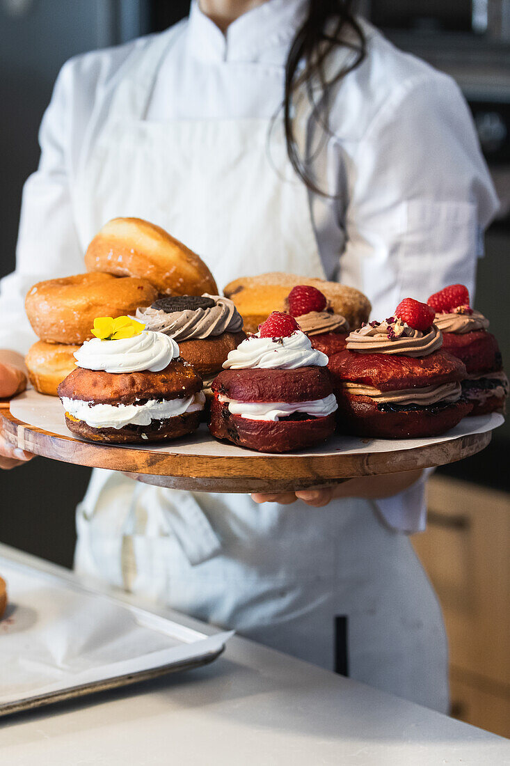 Crop anonymous female baker in uniform standing in bakery with tray of tasty vegan Berliners with whipped cream filling