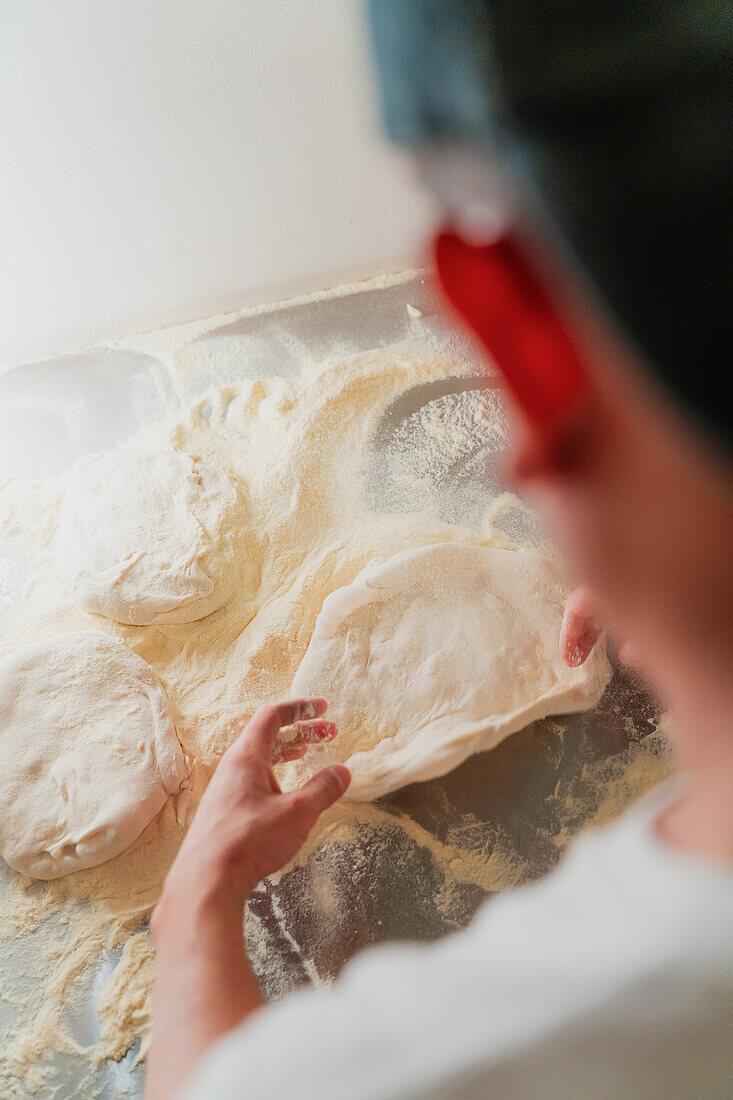 Ein Blick über die Schulter eines Pizzabäckers, der in einer Pizzeria den Teig ausbreitet, mit einer gesichtslosen und beschnittenen Darstellung, die sich auf die Hände und den Teig konzentriert