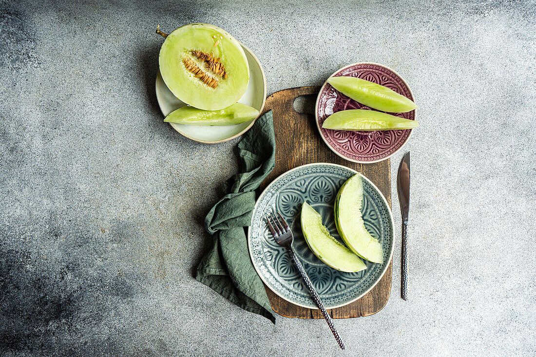 Draufsicht auf Bio-Mini-Melonenscheiben, die auf verzierten Tellern für ein erfrischendes Mittagessen angerichtet sind. Der rustikale graue Hintergrund unterstreicht das leuchtende Grün der frischen Melone.