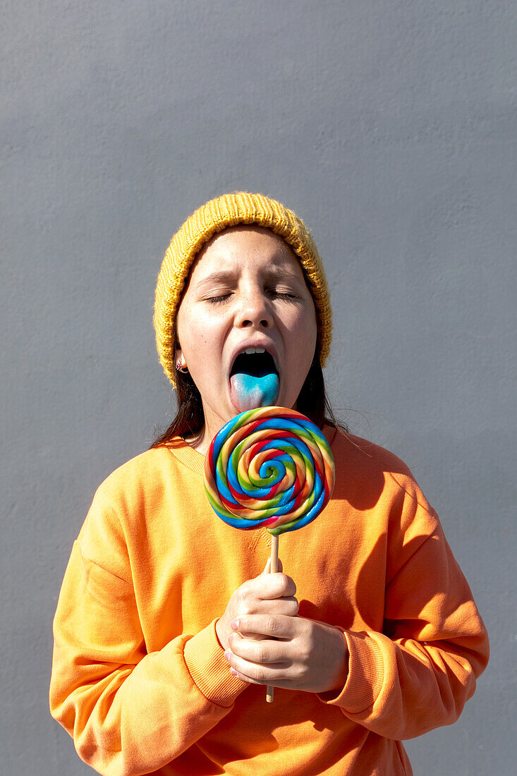 A young individual in an orange sweatshirt and yellow beanie savors the taste of a large, vibrant swirl lollipop against a plain background