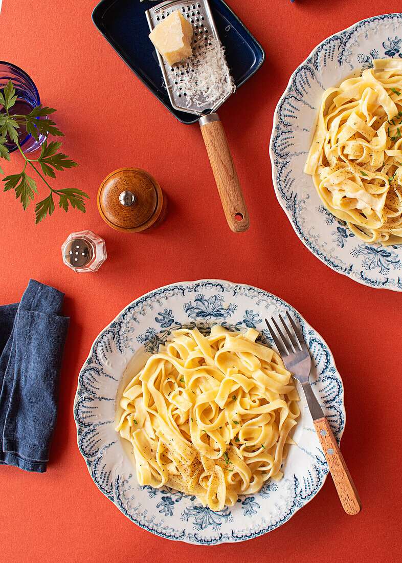 Von oben: Alfredo-Nudelgericht mit cremiger weißer Soße und Kräutern, serviert auf einem roten Tisch mit Pfeffermühle, Parmesanreibe und blauer Serviette