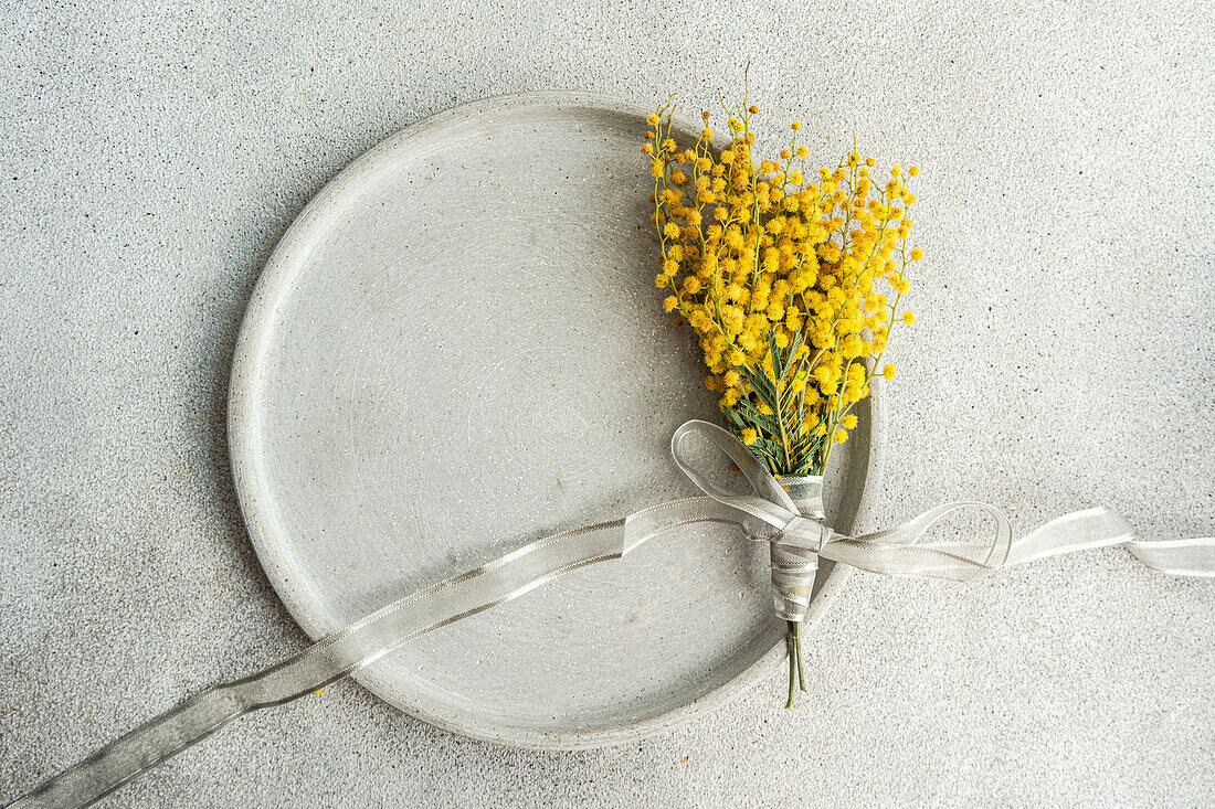 Draufsicht auf ein zartes Arrangement aus leuchtenden Mimosenblüten, die mit einem silbernen Band gebunden sind und auf einer glatten, strukturierten Tischoberfläche präsentiert werden, um einen raffinierten Akzent zu setzen