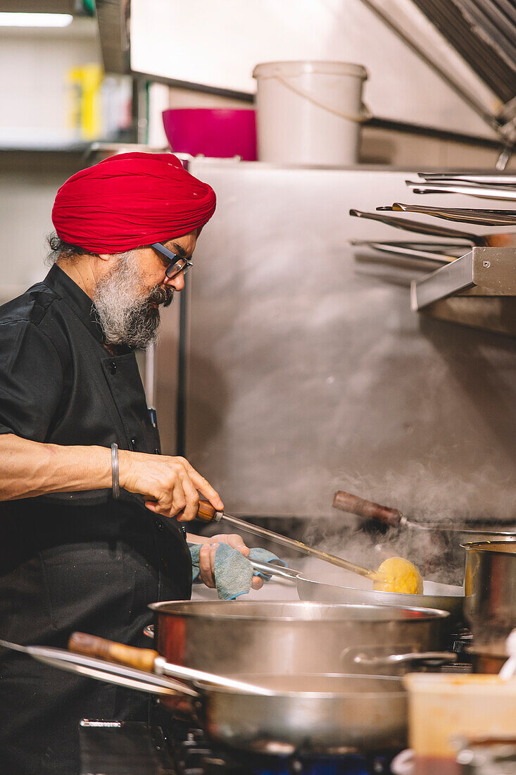 Ein Sikh-Koch mit rotem Turban kocht aufmerksam in einer geschäftigen Großküche, aus deren Töpfen Dampf aufsteigt