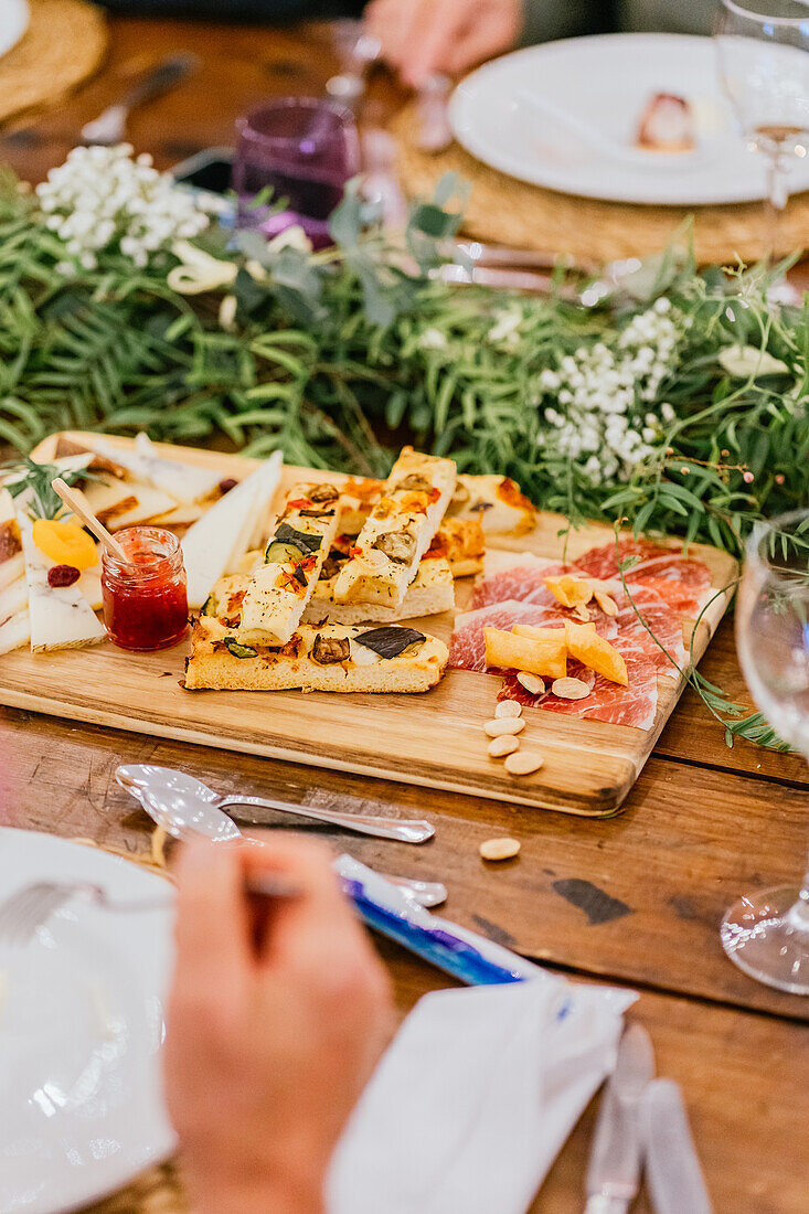 A rustic wooden board filled with a variety of cheeses, focaccia, and charcuterie, surrounded by fresh greenery and elegant tableware