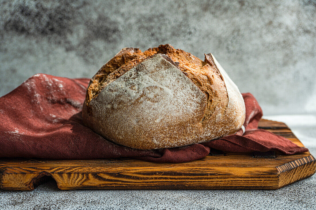 Freshly baked rye sourdough bread rests on a rustic wooden cutting board, draped with a burgundy cloth against a speckled grey background