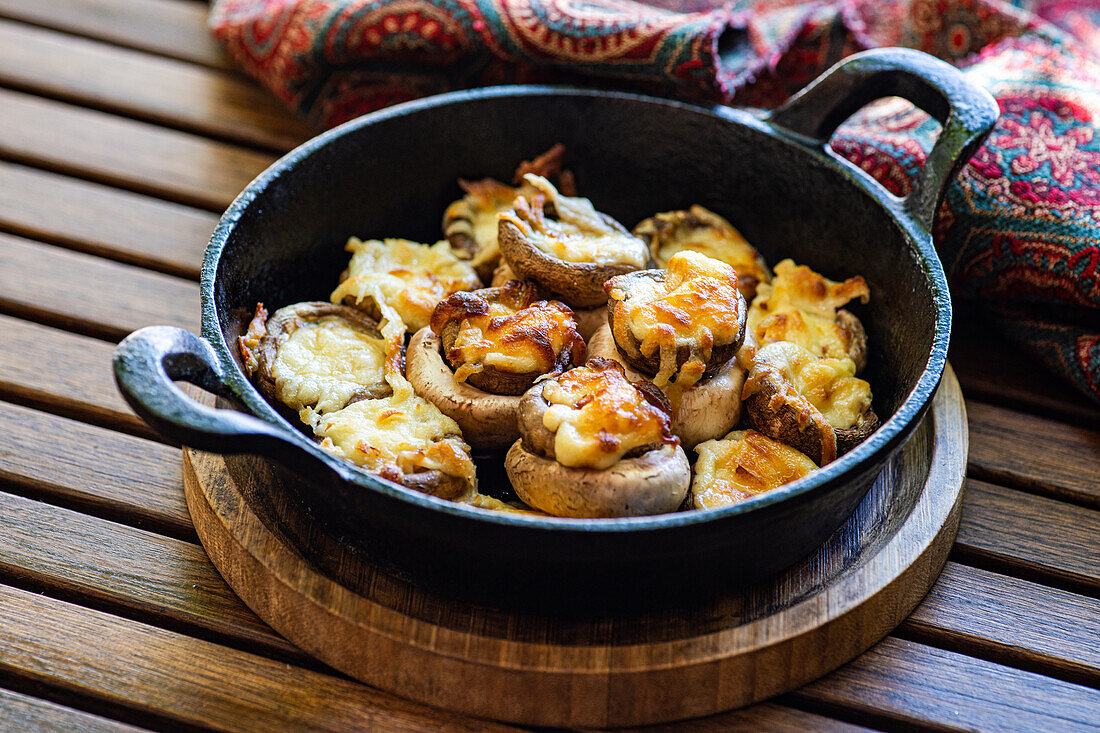 A tantalizing skillet full of baked mushroom caps generously topped with golden, melted Sulguni cheese, served on a wooden table, providing a rustic and appetizing visual