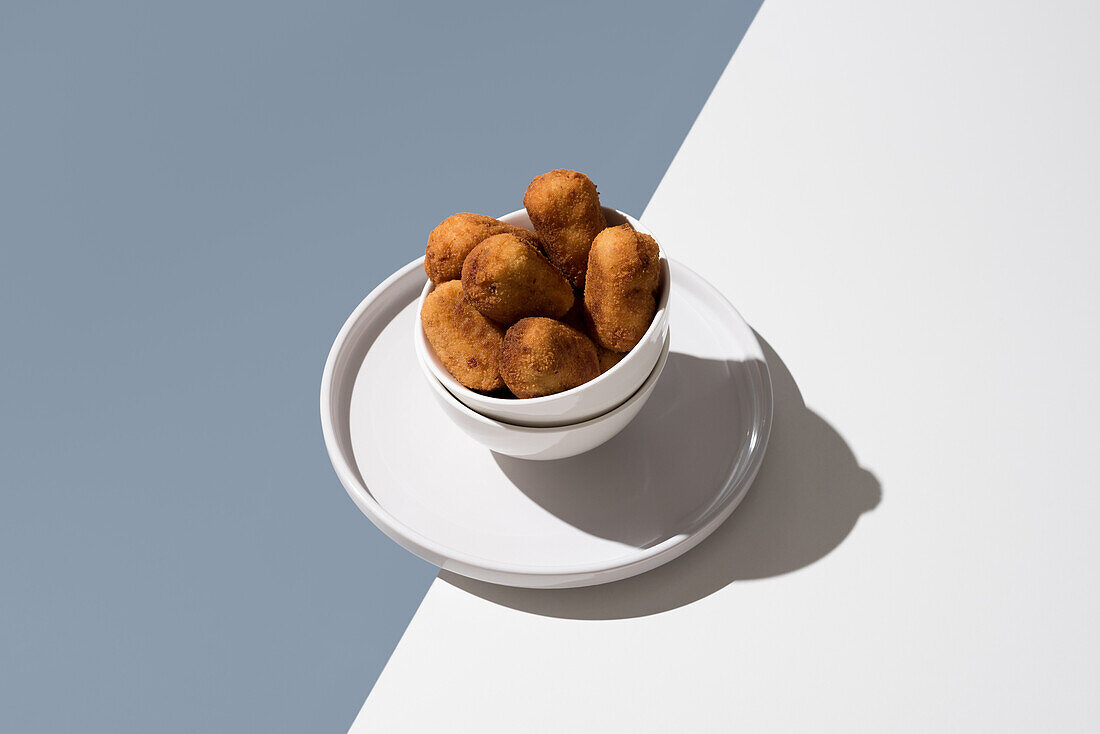 A minimalist composition featuring croquettes in a white bowl on a table, emphasized by natural shadows and a soft grey background