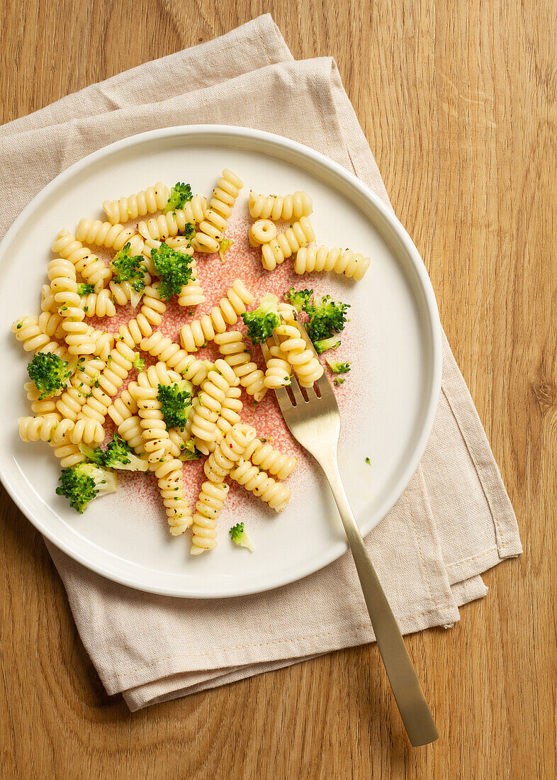 Elegant speckled plate holding a serving of fusilli pasta with broccoli florets, paired with a gold fork on a beige napkin and wooden background