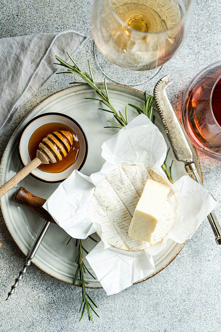 Draufsicht auf eine elegante Auslage von Brie-Käse mit Honig, Rosmarin, einem Korkenzieher und Gläsern mit Weiß- und Roséwein.