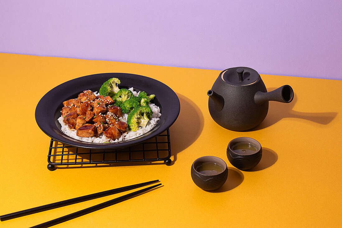 Artfully plated teriyaki chicken on a black plate, served with broccoli and rice, accompanied by a traditional tea set on a vibrant yellow and purple background.