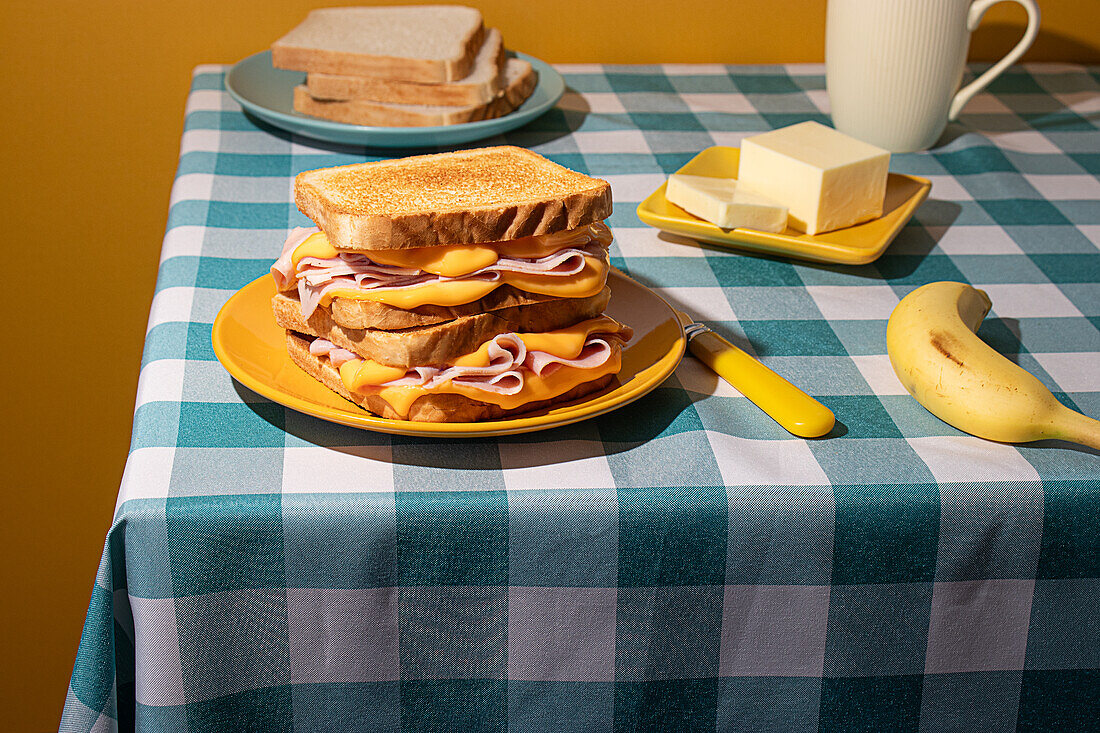 Ein hausgemachtes Frühstück auf einer karierten blauen Tischdecke mit einem Sandwich, einer Banane, Kaffee und Butter vor gelbem Hintergrund