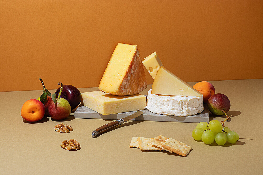 From above, an array of fine cheeses complemented by fresh fruit and nuts on a textured beige background, illuminated by soft lighting that accentuates their textures and colors