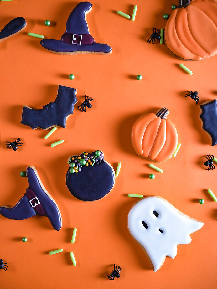 A festive assortment of Halloween cookies, artfully arranged on an orange surface The cookies, shaped like ghosts, witch hats, bats, cauldron and pumpkins, are iced in bright colors, accompanied by decorative spiders and candy sprinkles