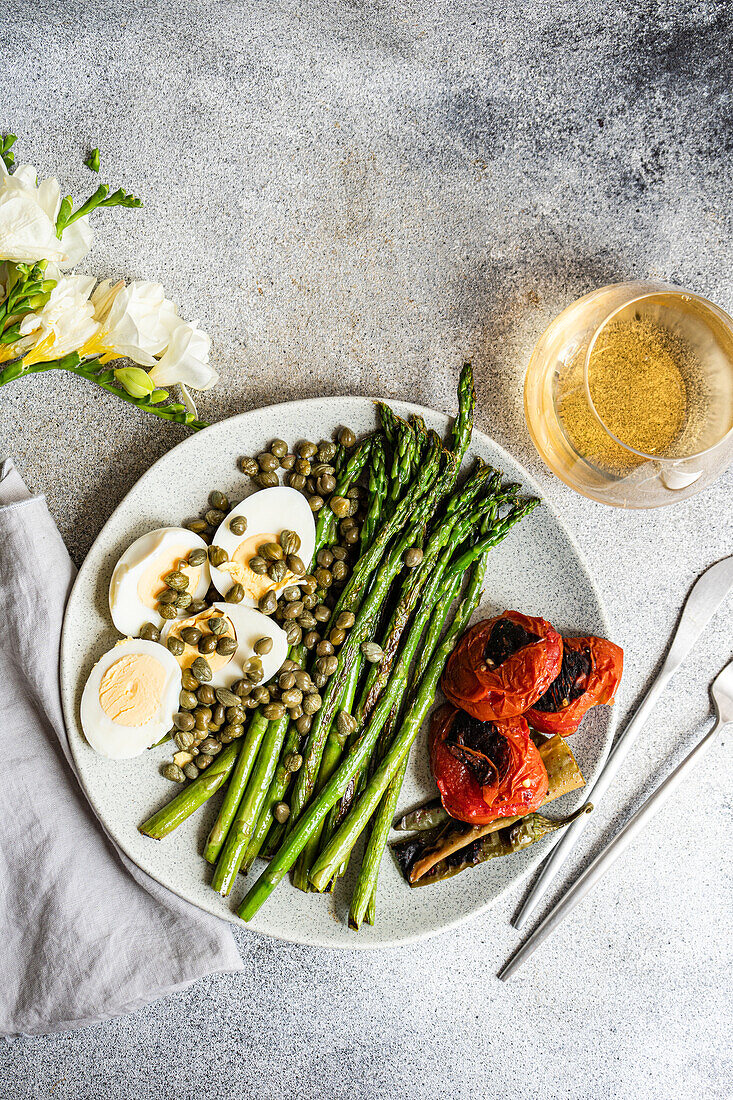 A neatly arranged plate showcasing healthy lunch options with a boiled egg, barbecued asparagus, capers, and brightly grilled vegetables including tomatoes and chili peppers, perfect for a nutritious meal.