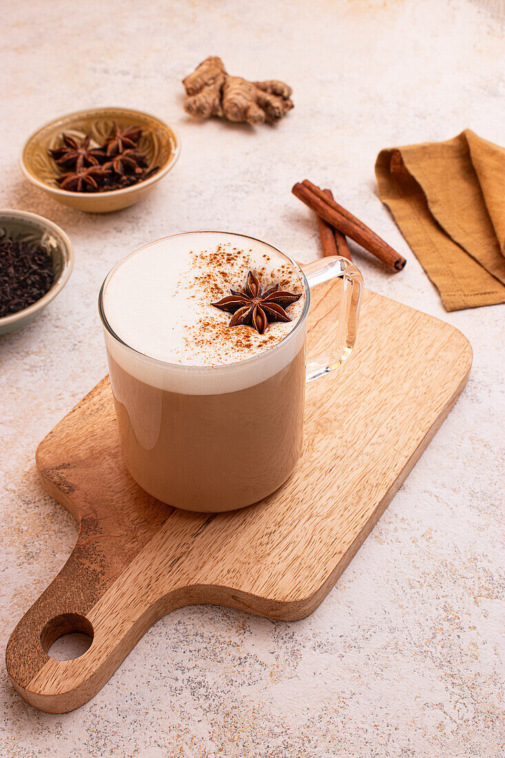 From above, Delightful chai latte with creamy foam on a wooden board surrounded by star anise, cinnamon sticks, fresh ginger, tea leaves, a milk jar and a brown napkin