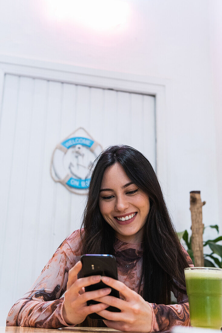 Positive female sitting at table with smoothie and browsing mobile phone while chilling at weekend in bar