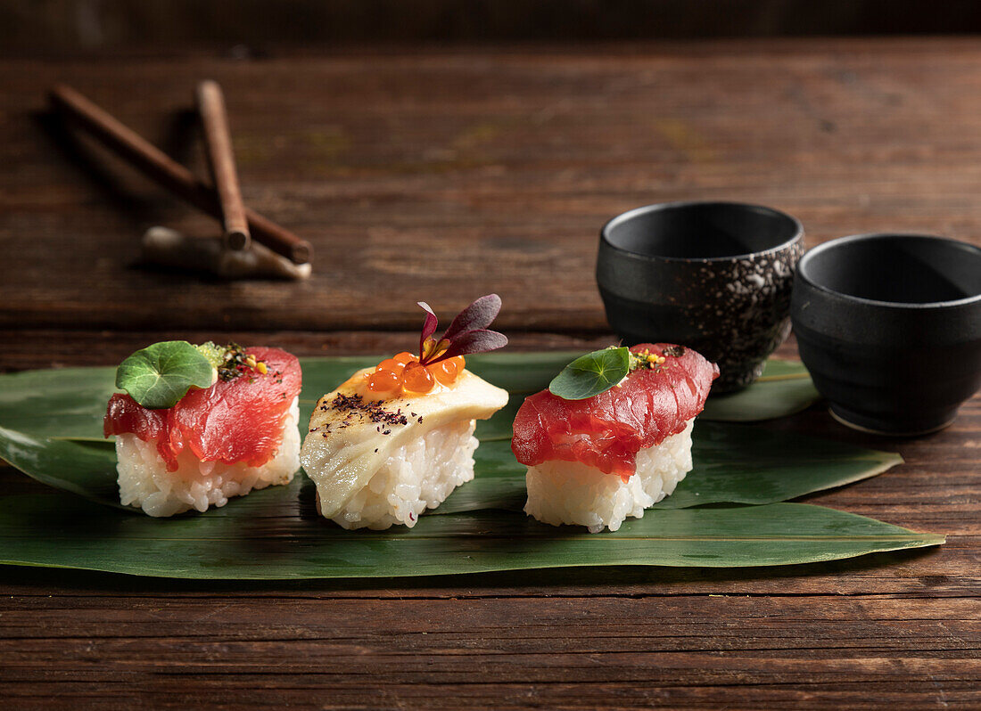 A trio of sushi pieces elegantly presented on a broad green leaf, accompanied by dark ceramic bowls and chopsticks, capturing the essence of Japanese culinary artistry