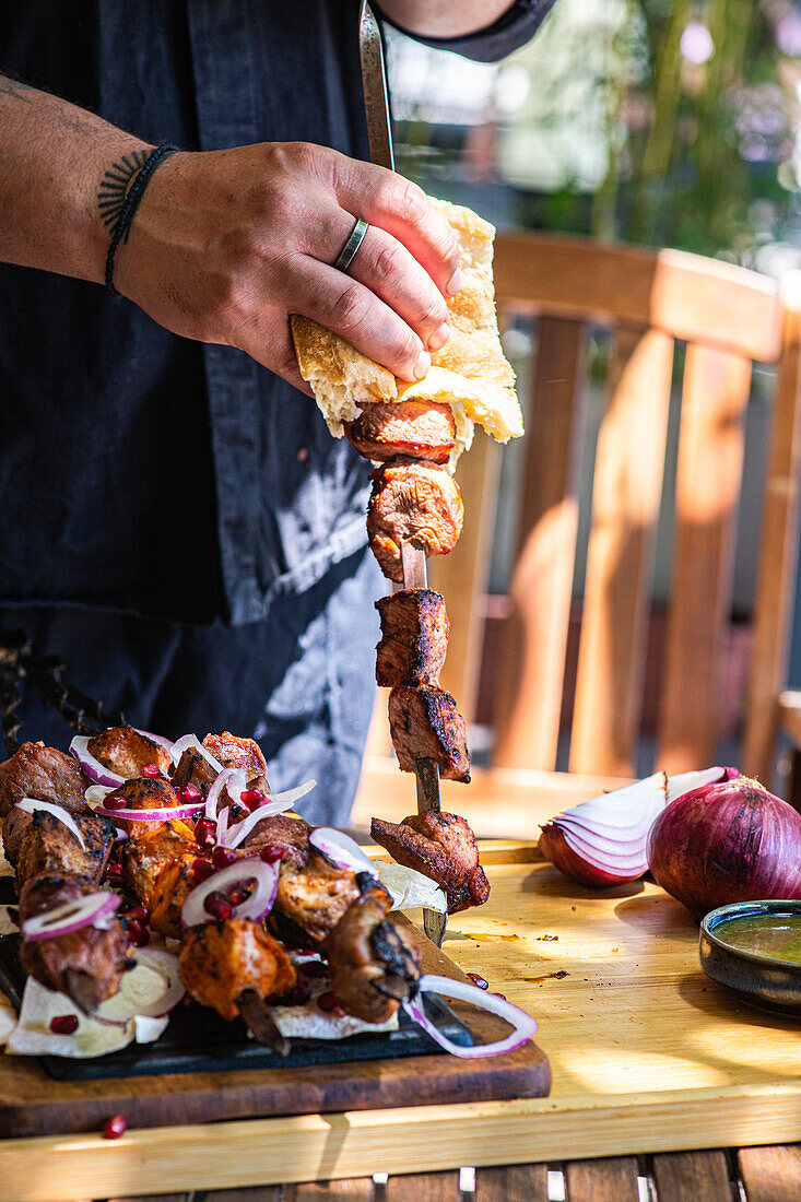 Eine lebhafte Aufnahme, die die Zubereitung des traditionellen georgischen Mzvadi zeigt, mit saftigen Fleischspießen, die an einem sonnigen Tag genossen werden Eine Person probiert das gebratene Fleisch, begleitet von Brot, geschnittenen Zwiebeln und einer Soße