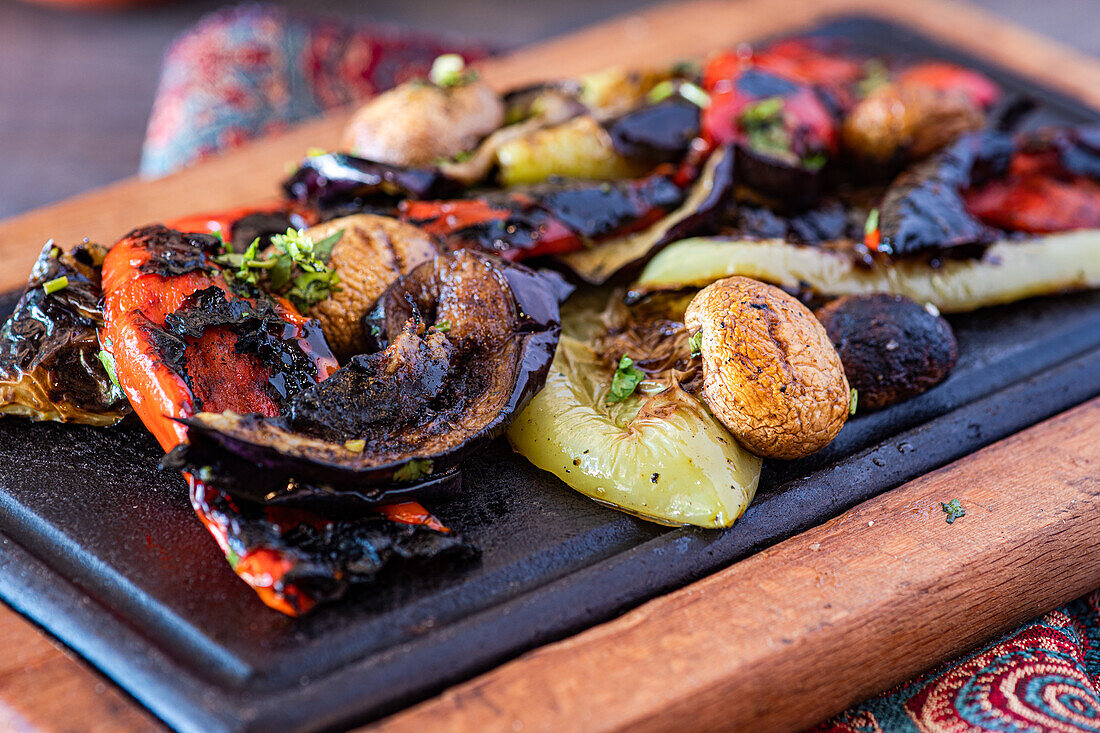 Eine bunte Auswahl an gegrilltem Gemüse, darunter Paprika, Pilze und Auberginen, präsentiert auf einem dunklen Holzbrett, gewürzt mit frischen Kräutern