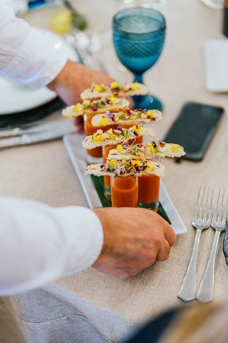 Ein anonymer Koch stellt Gourmetsuppen mit Crackerbelag auf einen elegant gedeckten Tisch, bereit für ein gehobenes Esserlebnis.