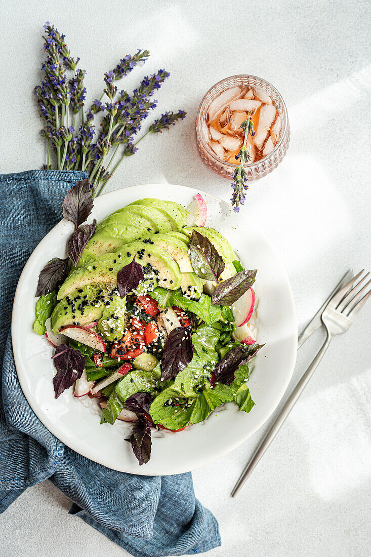 A vibrant vegetable salad with fresh lettuce, olives, cherry tomatoes, cucumber slices, radish, and red basil, topped with black and white sesame seeds, artfully presented in a white bowl.