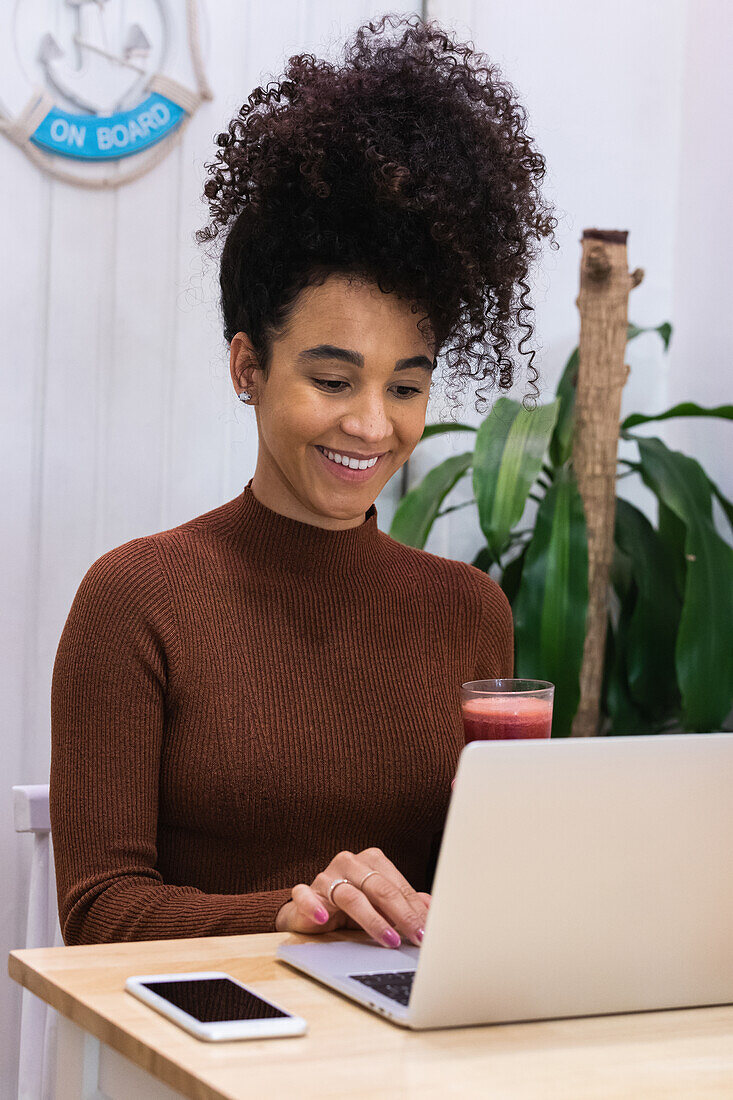 Fokussierte afroamerikanische Freiberuflerin, die mit einem erfrischenden Smoothie am Tisch sitzt und auf ihrem Netbook surft, während sie in einem Café an einem Projekt arbeitet