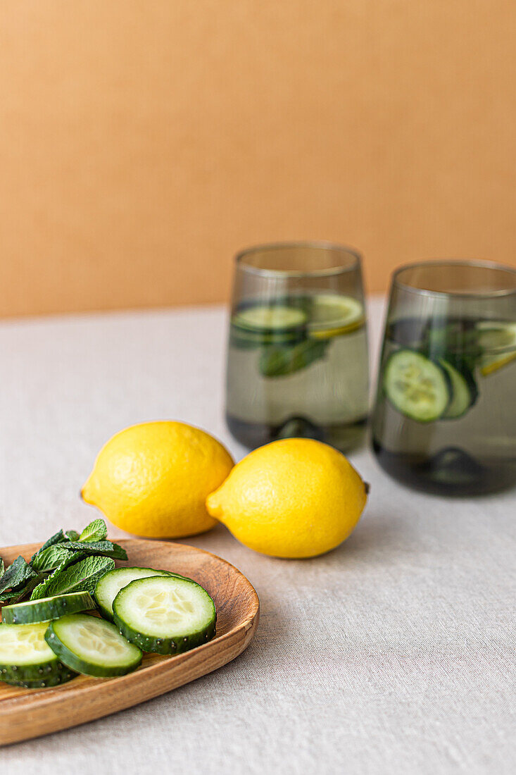 Zwei mit Gurken- und Zitronenwasser gefüllte Gläser, die mit frischen Zitronen und Gurkenscheiben auf einem Holztablett präsentiert werden und für eine gesunde Lebensweise werben.