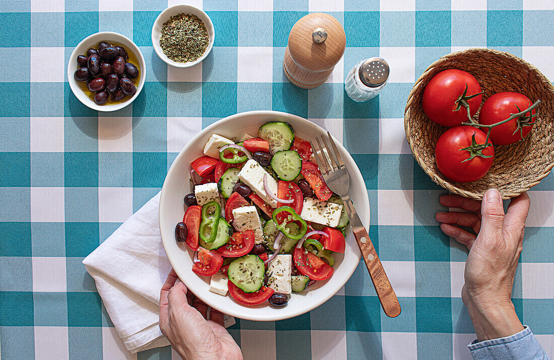 A freshly prepared Greek salad with tomato, cucumber, feta cheese, and olives, served on a blue and white checkered tablecloth