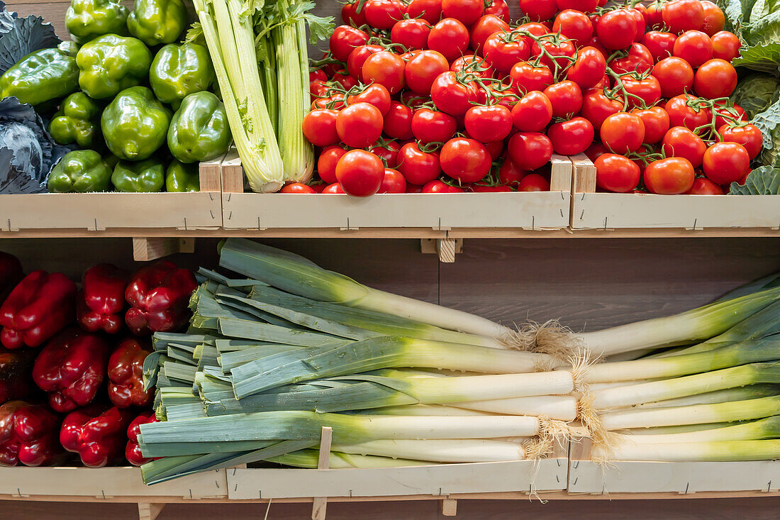 A variety of fresh vegetables, including green bell peppers, ripe tomatoes, red bell peppers, and leeks, neatly organized at a market stall